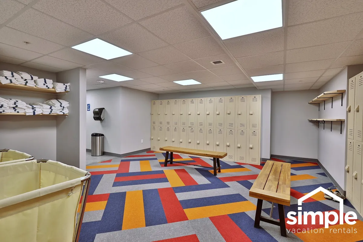 The scene shows colorful patterned flooring, storage lockers, wooden benches, and stacks of towels – a practical, organized changing area for guests coming from the pool or fitness center.