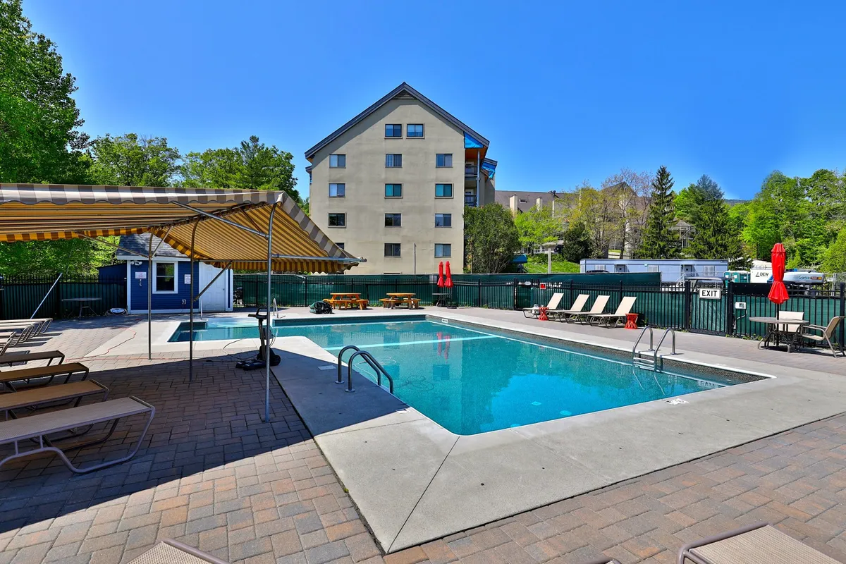 An inviting exterior pool area with sparkling blue water, plenty of lounge chairs, a striped awning for shade, and picnic tables nearby. Set against a lush, green backdrop, this outdoor space is perfect for summer swims, sunbathing, and casual barbecues.