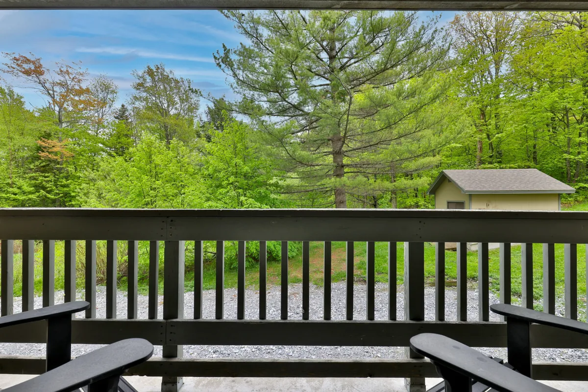A tranquil exterior scene featuring lush green trees, a gravel path, and a wooden railing, with a shed visible in the background. The porch area invites relaxation and offers views of the surrounding landscape, evoking a sense of connection to nature.