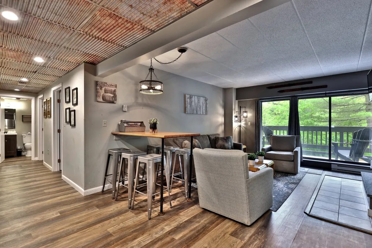 A stylish communal space featuring a wooden bar table and cozy seating area, with large windows offering views of greenery. The space is bathed in natural light, perfect for socializing, relaxing, or planning the evening ahead.