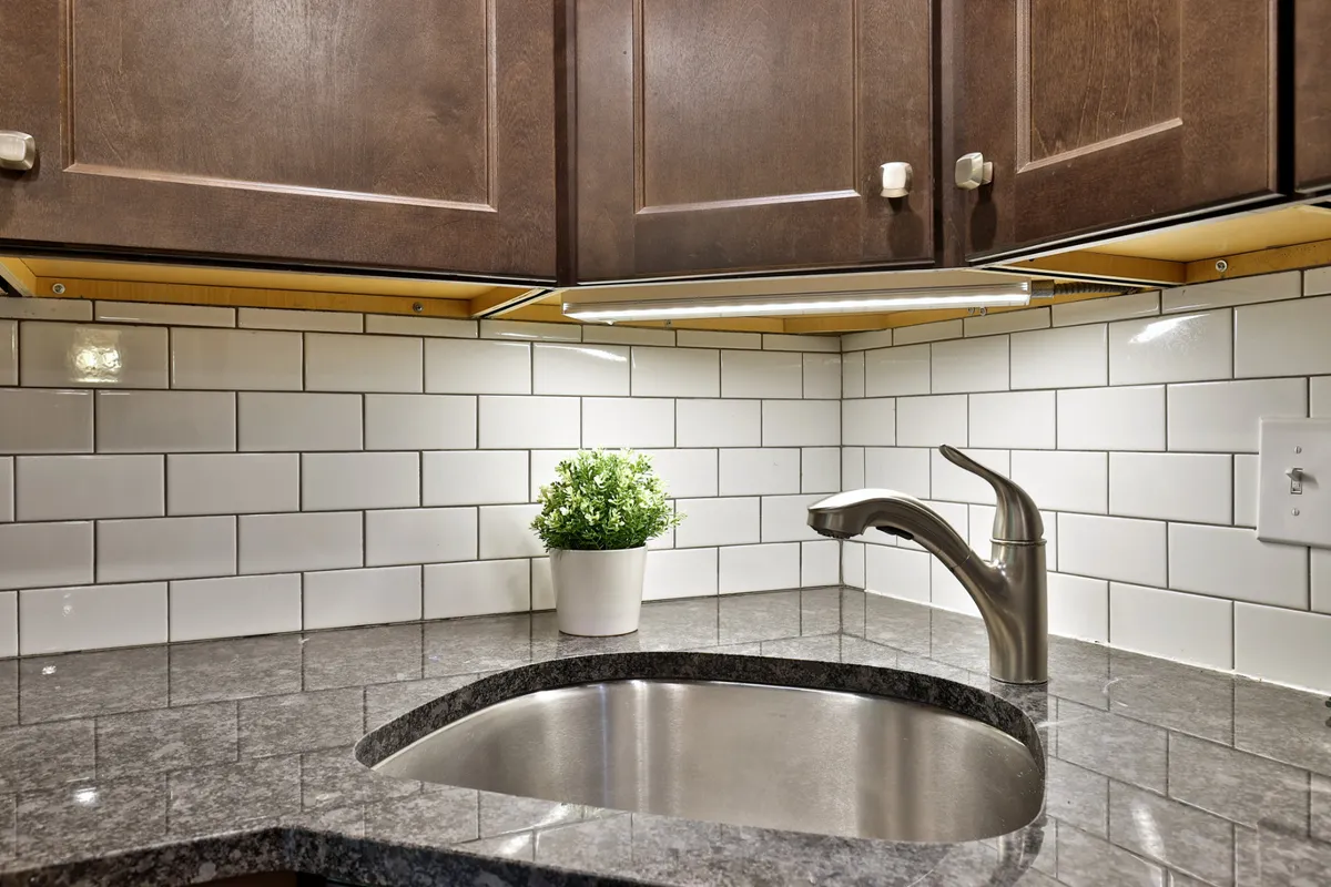 A bright kitchen space featuring a granite countertop, modern faucet, potted plant, and subway tile backsplash. The scene conveys opportunities for self-catering, meal prep, and social gatherings, all of which help stretch guest budgets.