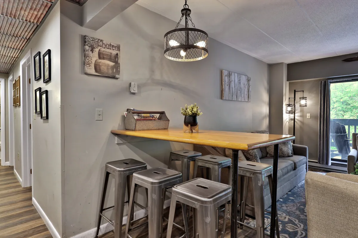 A cozy dining area featuring a modern wooden table with metal bar stools, a charming light fixture overhead, decorative wall art, and greenery in a vase. The space feels inviting for casual dining, morning rituals, or celebratory meals.