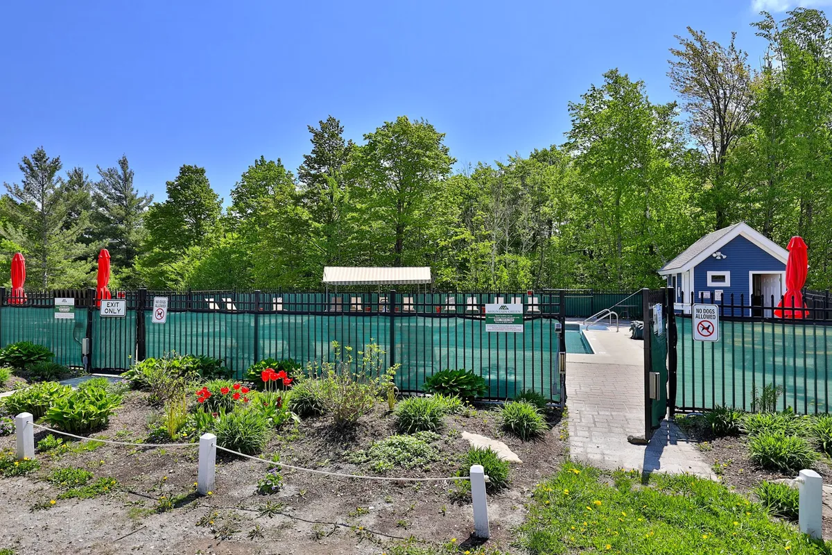 An inviting outdoor swimming pool surrounded by red umbrellas, lush green foliage, and vibrant flower beds. The scene features guests lounging poolside, enjoying the water, and soaking up the sun in a cheerful, communal atmosphere.