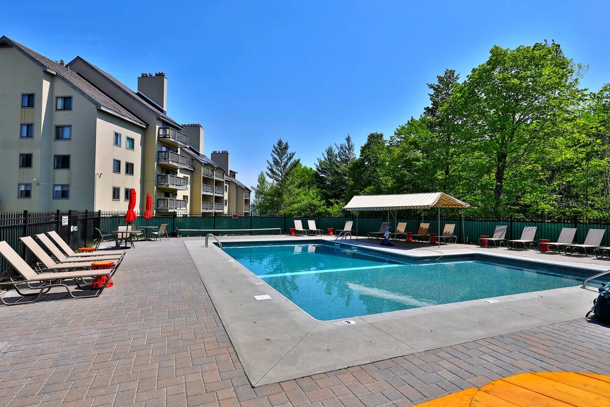 A bright exterior shot of the swimming pool area, with lounge chairs and green trees bordering the space. The building’s exterior is visible, suggesting easy access to outdoor amenities, while groups of friends and families enjoy the refreshing water and the sun.