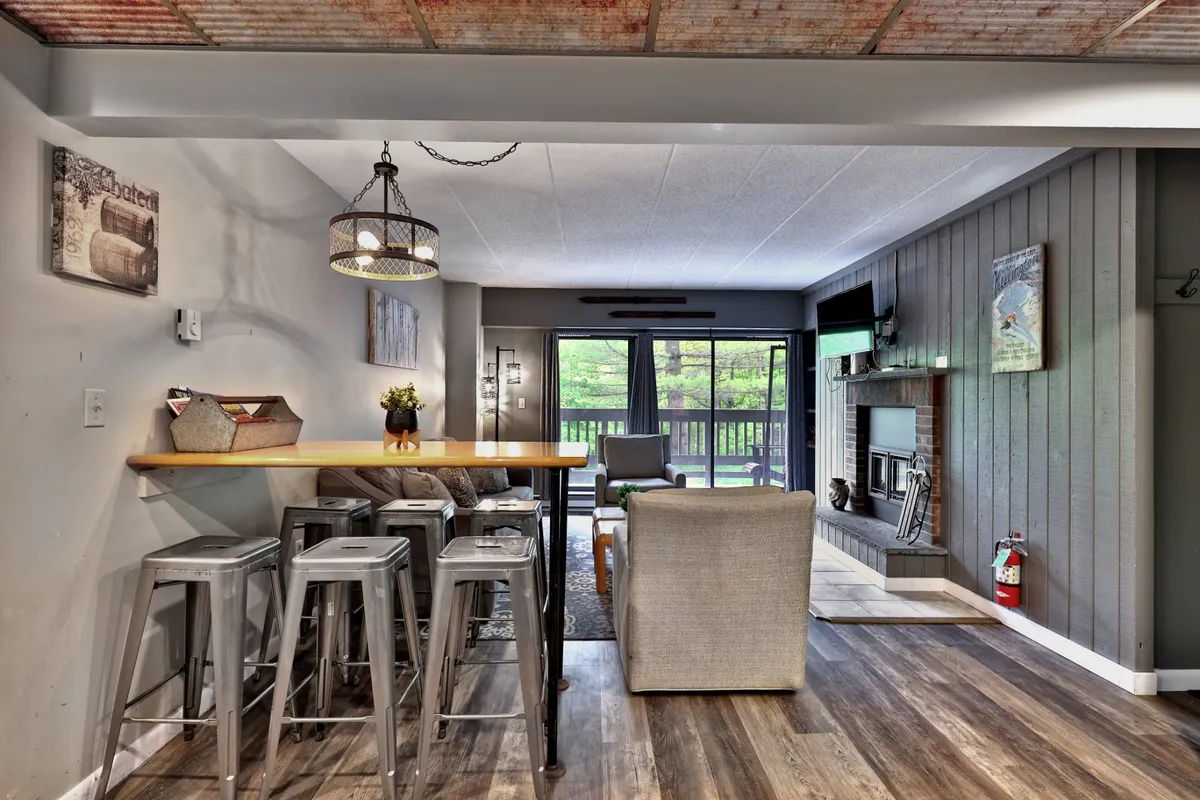 A bright and open living space featuring a bar counter, fireplace, and expansive windows that lead out to an outdoor deck or balcony. The setup invites gatherings, whether for casual dining or cozying up by the fire, and offers a seamless transition between indoor comfort and outdoor enjoyment.