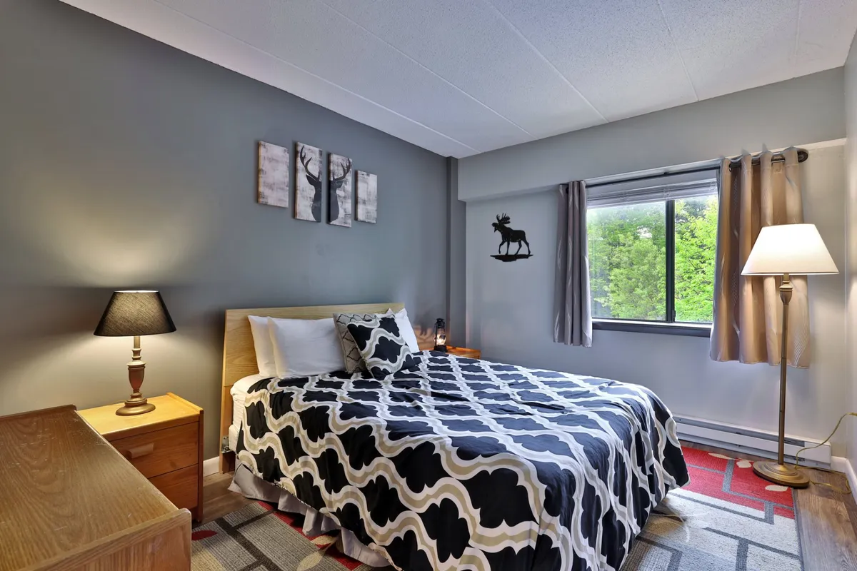 A cozy bedroom with a bed adorned in patterned bedding, a nightstand with a lamp, a window letting in natural light, and decorative wall art. The space feels inviting, tranquil, and designed for rest.