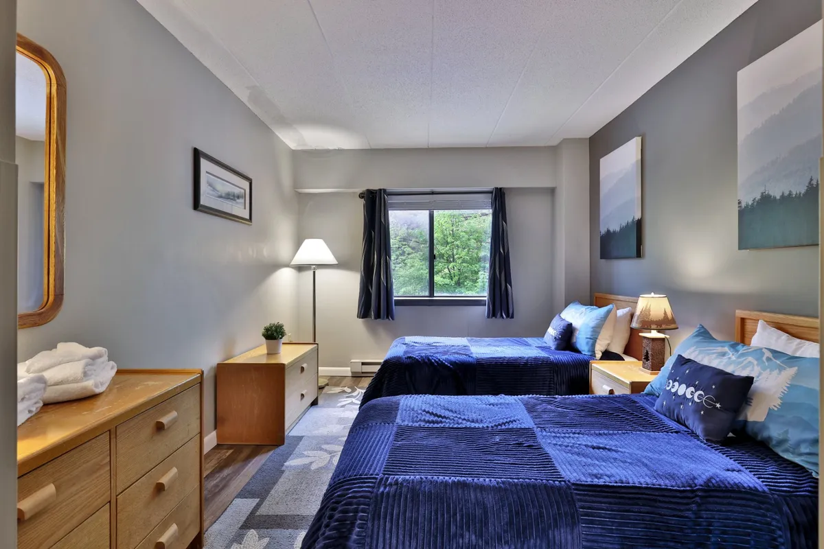 A bedroom with twin beds, a window view, a nightstand with a lamp, decorative mirror, and wall artwork. The setting looks comfortable and perfect for kids or friends, offering flexibility in sleeping arrangements.