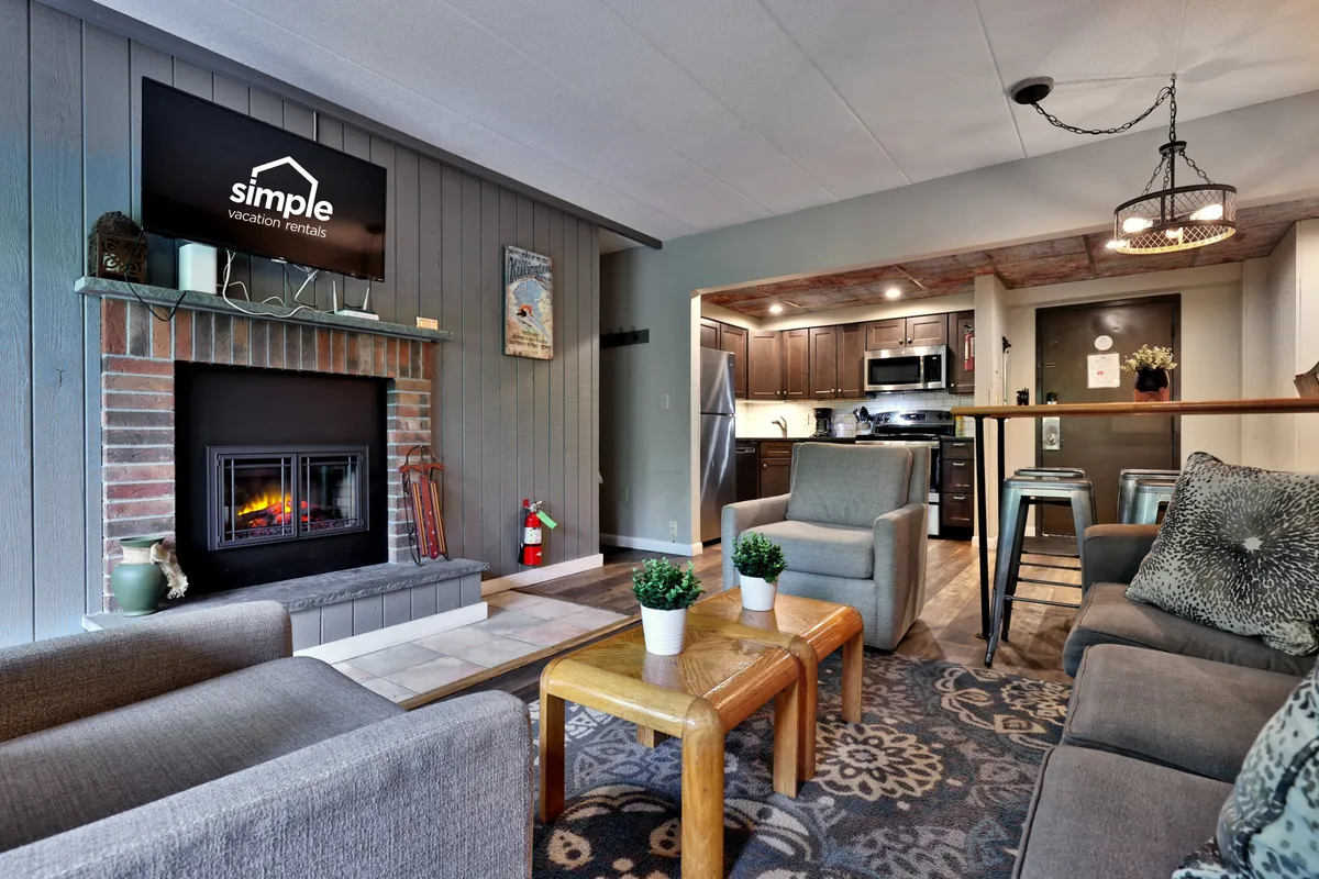 This image showcases a modern living room with a stylish fireplace, comfortable seating, and a view of the kitchen in the background. Large windows allow natural light to fill the space, and a wall-mounted TV is visible, perfect for movie nights and family gatherings.