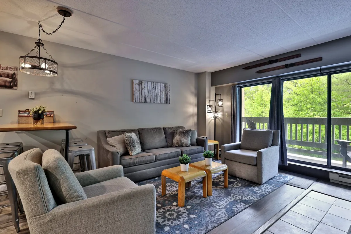 The image showcases a cozy living area with a plush sofa and coffee table, set beside a large window that frames a tranquil mountain view. Natural light fills the room, highlighting the inviting space perfect for unwinding after a day outdoors.